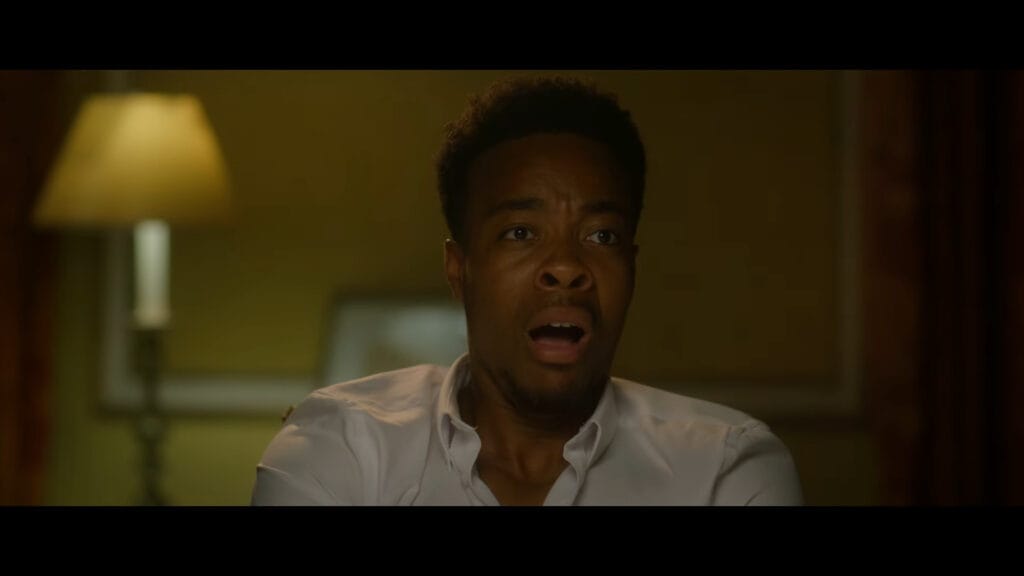 A close-up shot of a young Black man wearing a white shirt looking completely shocked and terrified with his mouth wide open in a dimly lit room.