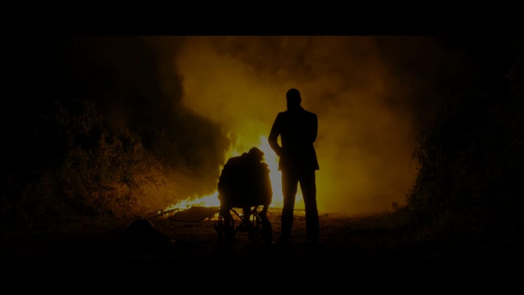 A dark silhouette of a man standing next to someone in a wheelchair, backfacing the camera against a massive, roaring wall of fire in the background.