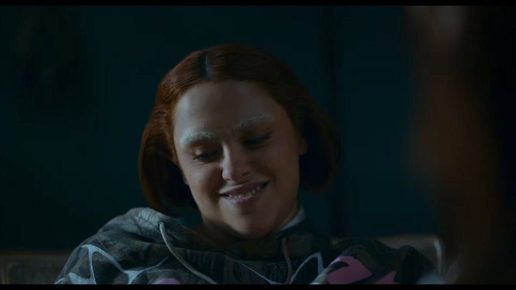A close-up shot of a smiling woman with short reddish-brown hair and bleached eyebrows wearing a camouflage hoodie in a scene from No Me Sigas.