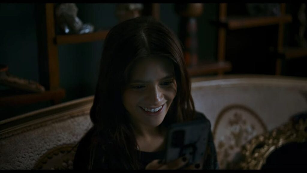 Carla smiling brightly as she looks down at her mobile phone screen in a dimly lit room in the social media horror movie No Me Sigas.
