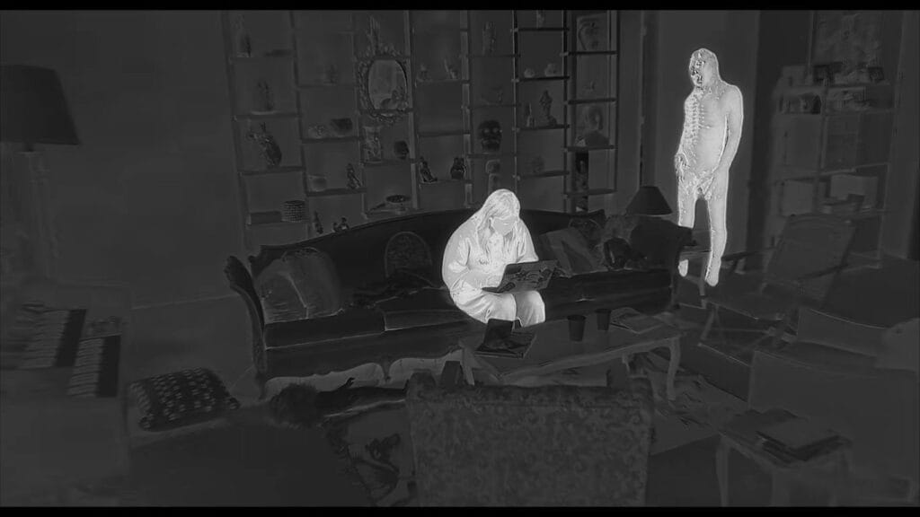 A chilling thermal or night-vision style shot from No Me Sigas showing a glowing figure on a couch with a laptop as a tall, skeletal apparition stands menacingly behind her.
