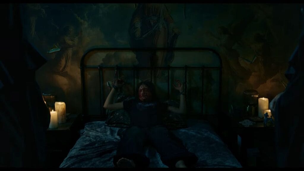 Carla tied to a metal bed frame with blood covering her face in a dark room lit by candles under a large painting from a dramatic scene in No Me Sigas.