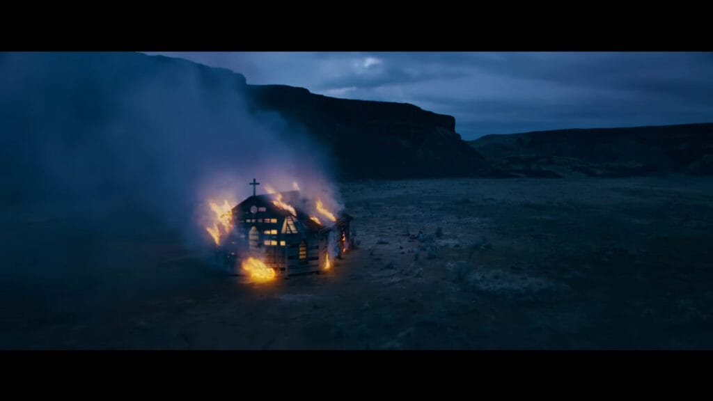 An aerial shot of a small, isolated wooden church engulfed in bright flames at twilight, a central visual element for a Passenger ending explained discussion.