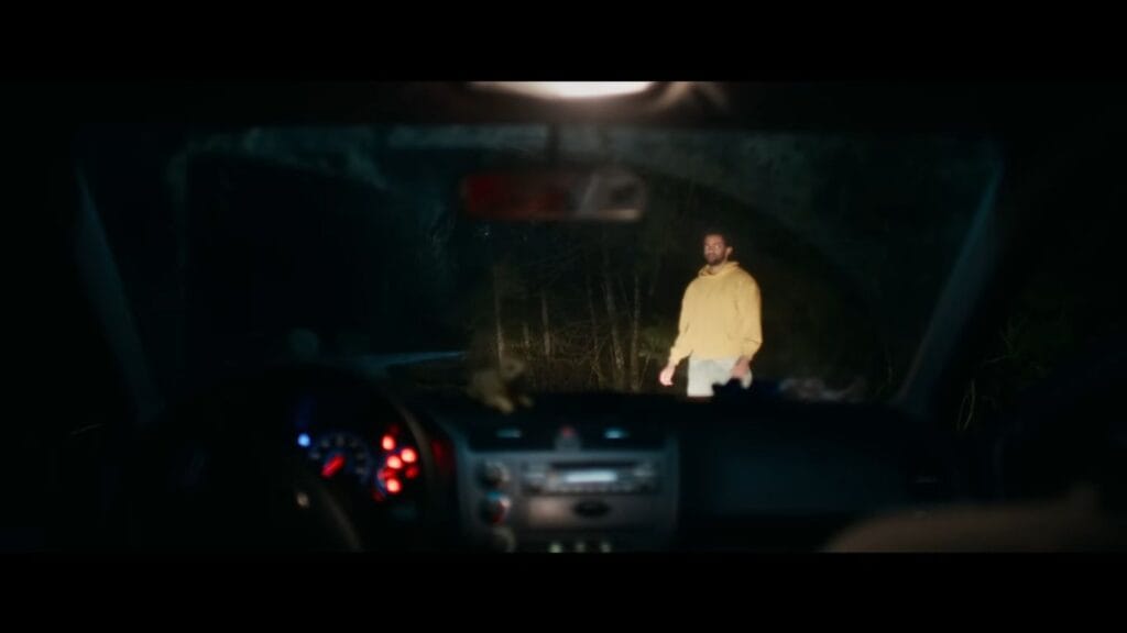A view from inside a dark car looking through the front windscreen at a man wearing a yellow hoodie standing on a dark, tree-lined road at night, illuminated by the vehicle's headlights.