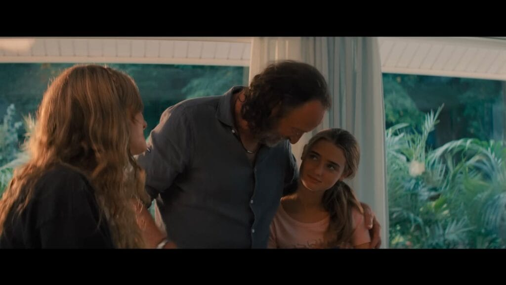 A father smiling and putting his arms around two young women inside a bright, sunlit room in a scene from the creature feature Primate.