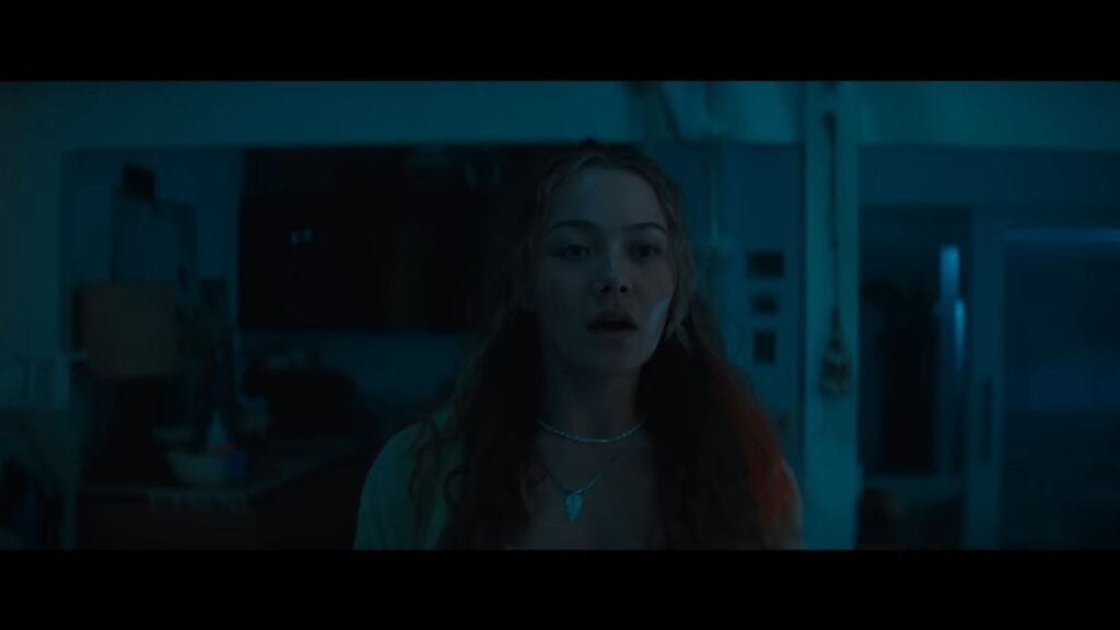 A young woman looking shocked and fearful in a dark blue, shadowy room in a tense scene from Johannes Roberts's Primate.
