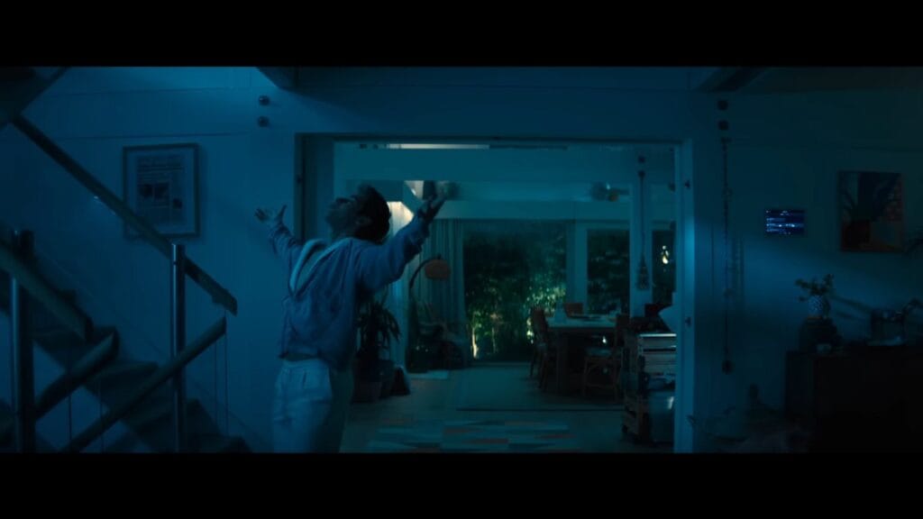 A man standing with his arms outstretched inside a dimly lit, modern cliffside home at night in a scene from the horror movie Primate.