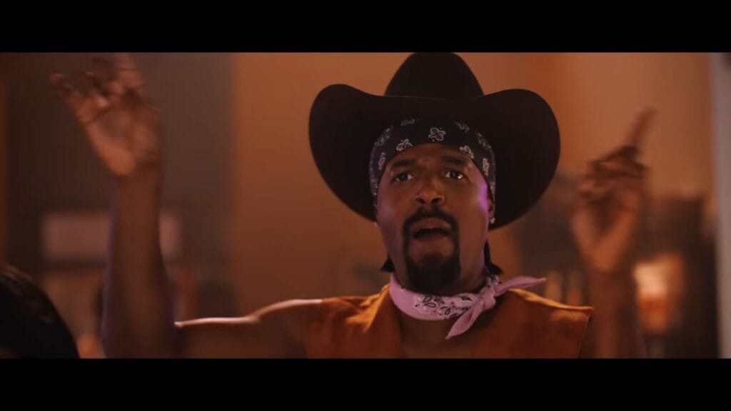 Shawn Wayans dressed in a cowboy hat, bandana, and leather vest dancing enthusiastically in a scene from Scary Movie.