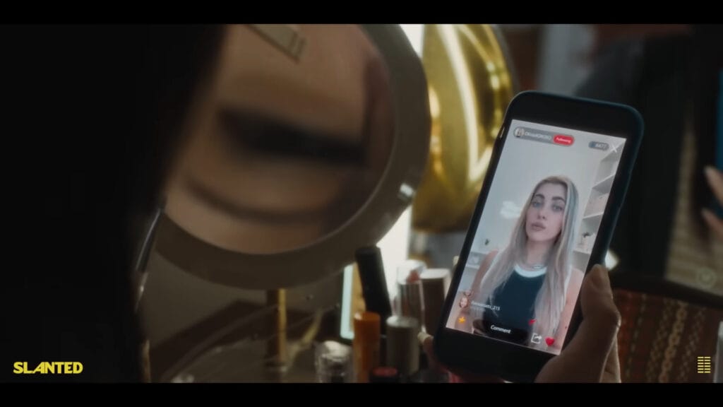 A first-person perspective shot from the film Slanted showing a character holding up a mobile phone displaying a blonde social media influencer stream next to a vanity mirror.