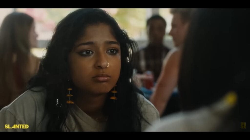 A close-up shot of a teenage girl with dark curly hair and a nose ring looking dejected and thoughtful in a busy school cafeteria scene from Slanted.