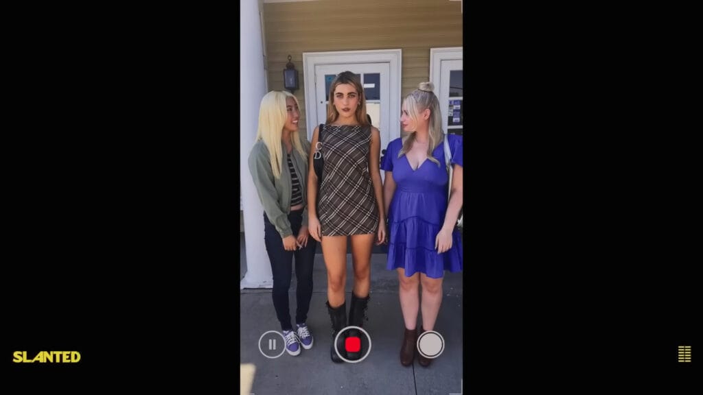 A smartphone video recording view showing three teenage girls posing together in trendy outfits outside a school building in a scene from Amy Wang's Slanted.
