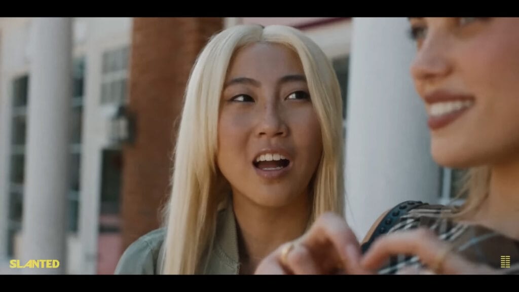 A close-up shot of a smiling teenage girl with bleached blonde hair talking to her friend outside in a scene from the dark comedy drama Slanted.