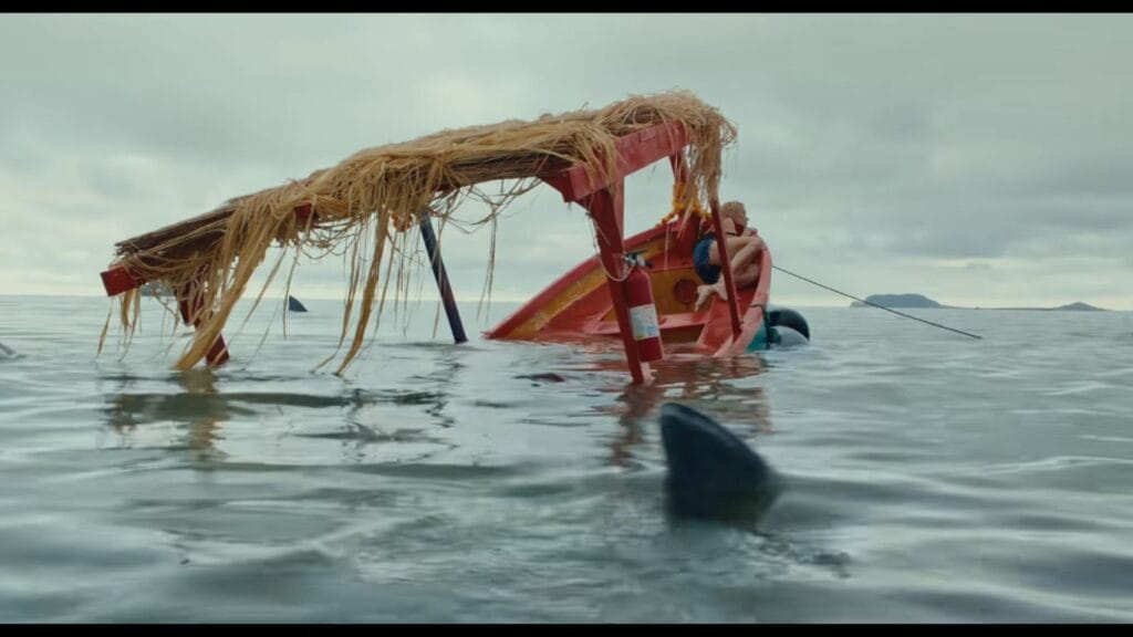 A small, red wooden tour boat taking on water and listing heavily in ocean waters surrounded by shark fins in a tense scene from The Bay.