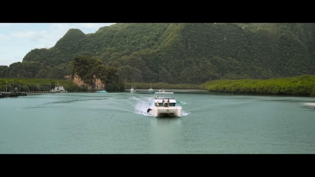 A white catamaran boat cruising through a wide green river surrounded by lush tropical mountains in Thailand in The Devil's Mouth.