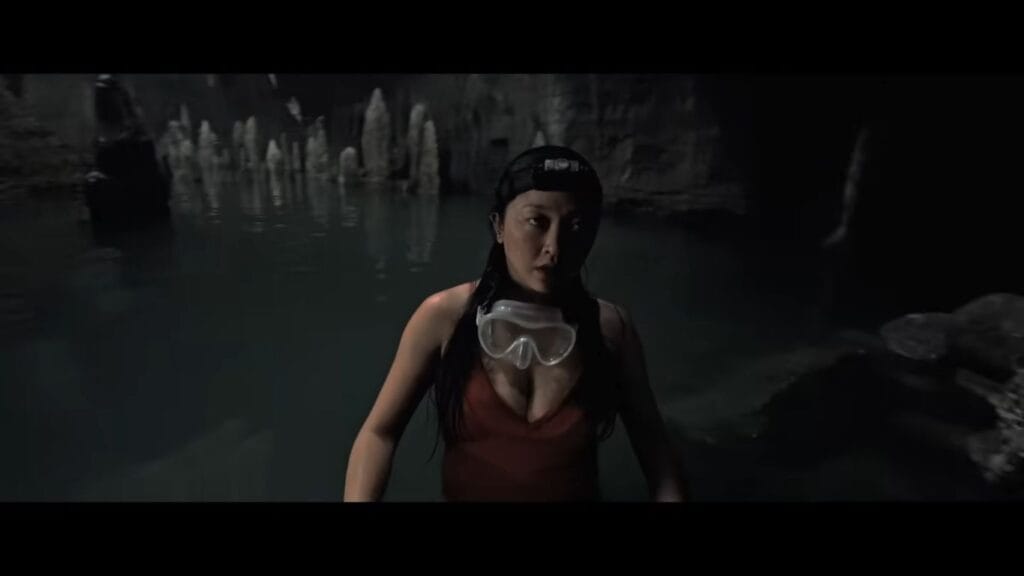 Lana Condor as Max standing chest-deep in water wearing a headlamp inside a shadowy cave system in The Devil's Mouth.