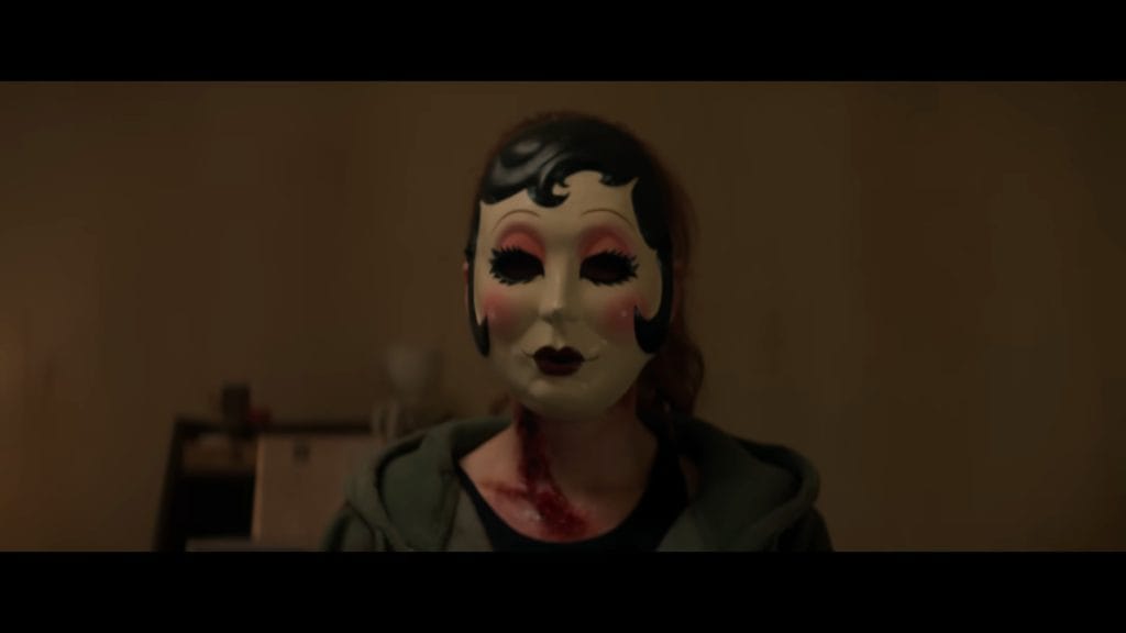 A person with a bloody neck injury wearing the iconic Pin-Up Girl doll mask and a green hoodie in a scene from The Strangers: Chapter 3.
