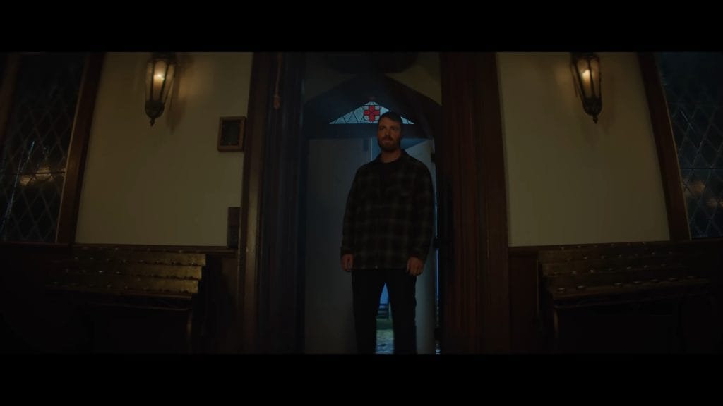 A man wearing a dark plaid shirt standing framed in a doorway under hanging lamps in a dim, church-like interior from Renny Harlin's The Strangers: Chapter 3.