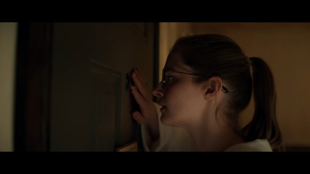A side-profile shot of a woman with a ponytail and glasses peering cautiously through a front door peephole in a scene from The Strangers: Chapter 3.