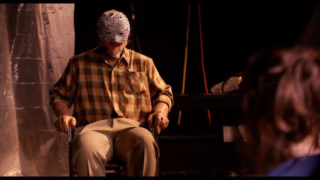 A man wearing a googly-eyed textured mask and plaid shirt seated in a chair facing a captive in Velvicide.