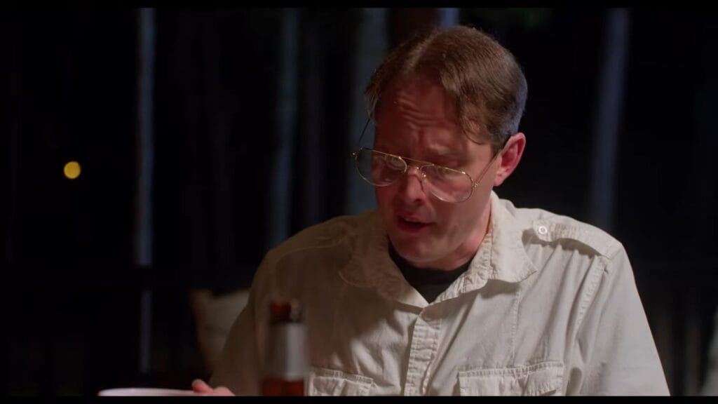 Jon Devlin wearing large wire-rimmed glasses and a white button-up shirt looking down in a scene from Velvicide.