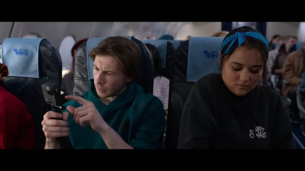A young man checking a small handheld action camera while sitting next to a passenger in an airplane in Black Box.