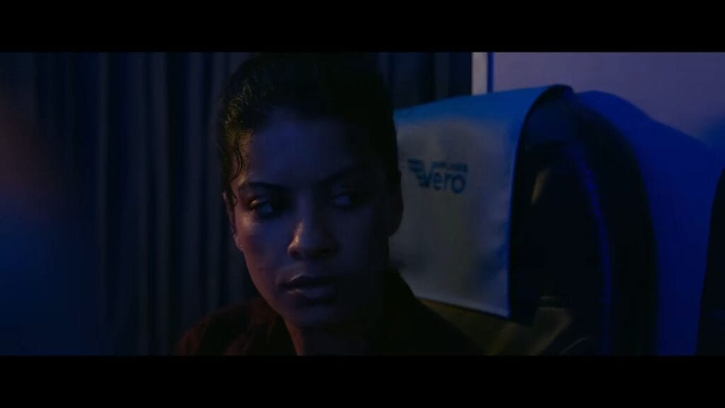 A woman in an airplane seat looking over her shoulder with an uneasy expression in dark blue lighting in Black Box.