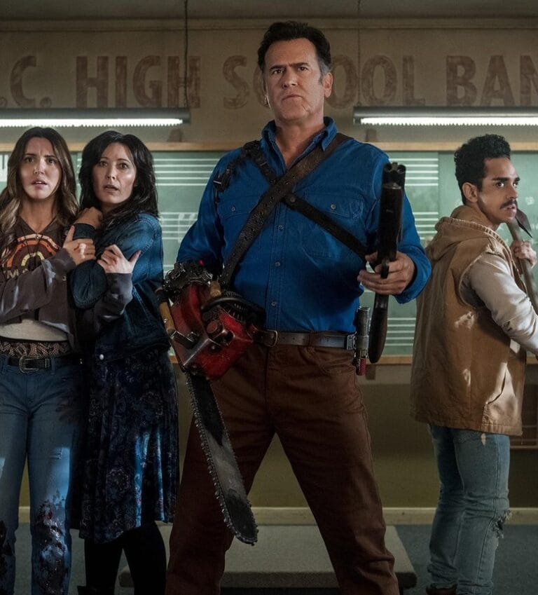 Bruce Campbell as Ash Williams holding a shotgun and chainsaw hand flanked by cast members in a classroom in Ash vs Evil Dead.