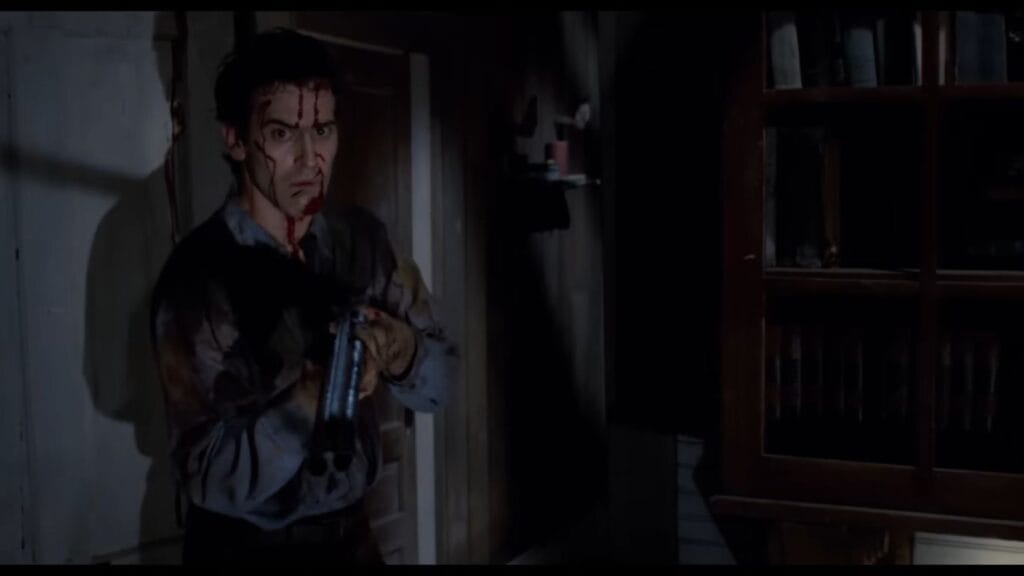 Bruce Campbell as Ash Williams covered in blood and aiming a shotgun with a crazed expression inside the cabin in Evil Dead II.