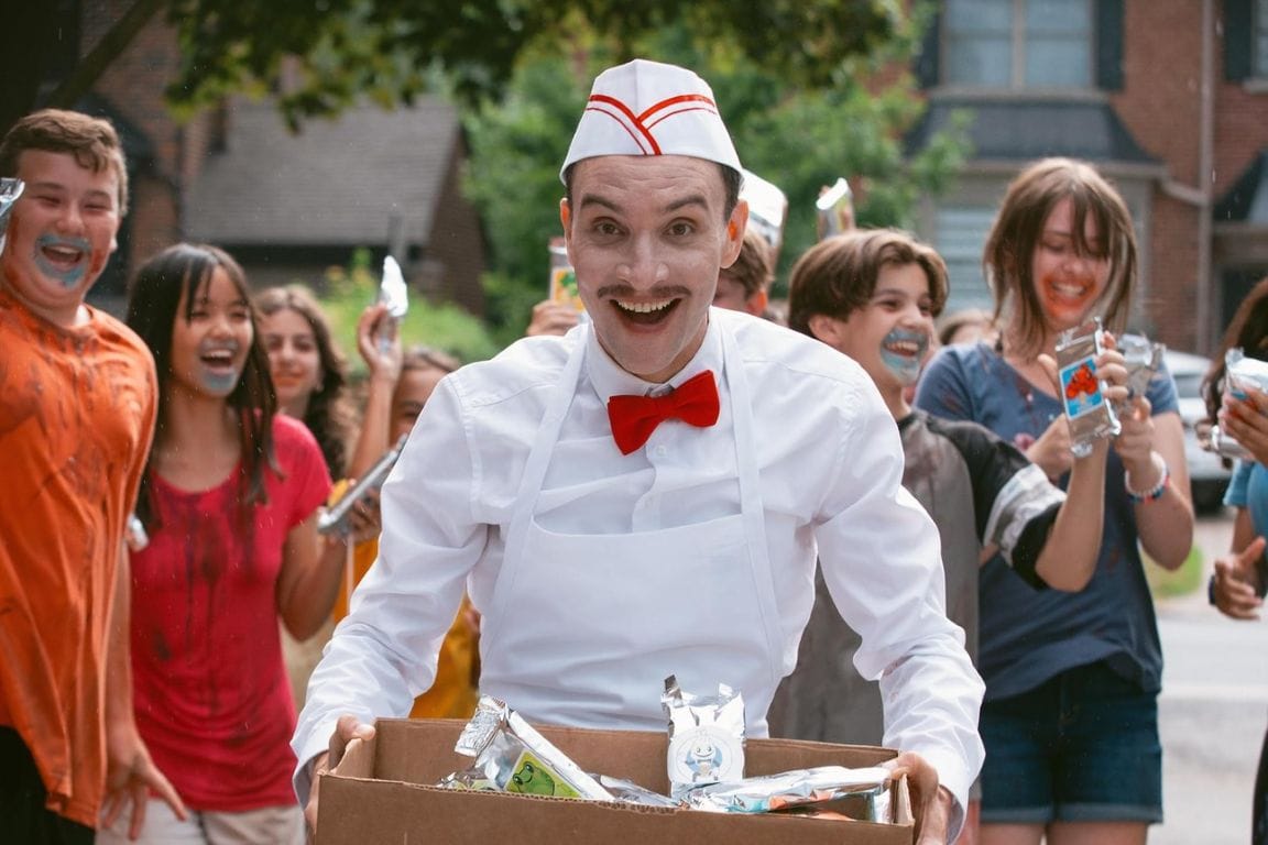 An ice cream vendor smiling maniacally while carrying a cardboard box full of treats as messy, excited children swarm behind him in Ice Cream Man.