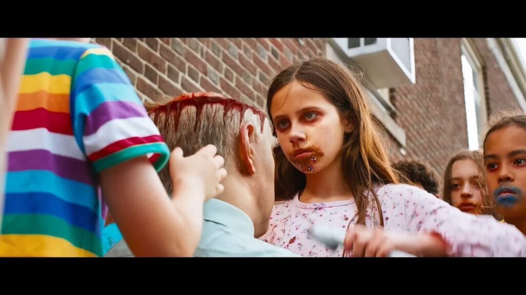 A young girl with chocolate and sprinkles around her mouth staring ominously at a man bleeding from the head as children gather around in Ice Cream Man.