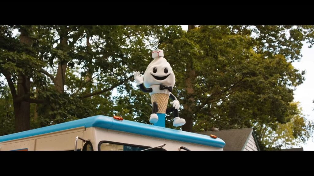 A large soft-serve ice cream mascot figure with a bow tie and paper hat waving from the roof of an ice cream truck in Ice Cream Man.