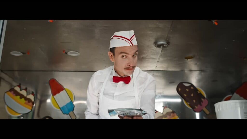 Ari Millen as the ice cream man wearing a white uniform and red bow tie holding out a packaged treat from his truck in Ice Cream Man.