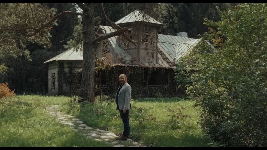 A bearded man in a blazer standing on a garden stone path in front of a derelict wooden house surrounded by dense Finnish forest in Nightborn.