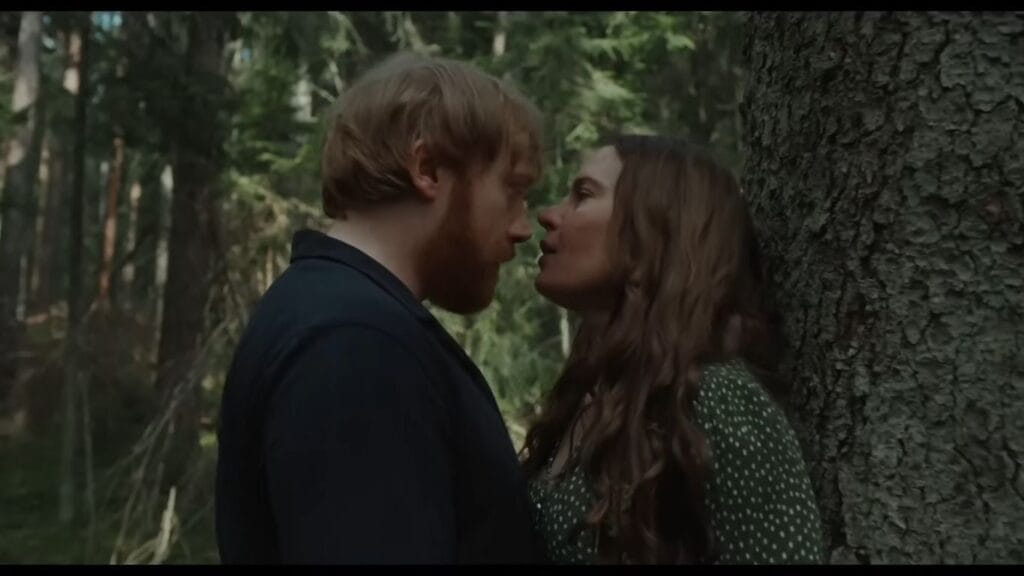 Saga leaning against a mossy tree trunk while intimately facing Jon in the deep woods in a scene from Nightborn.