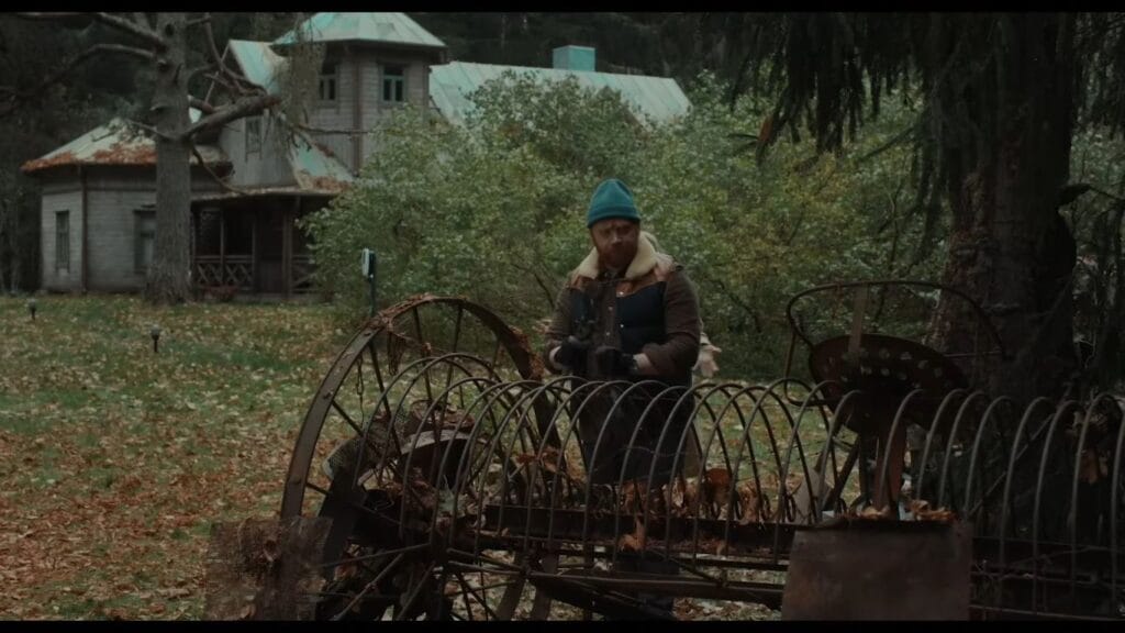 A man in a beanie and vest dismantling rusted farm machinery and scrap iron in the overgrown garden in Nightborn.