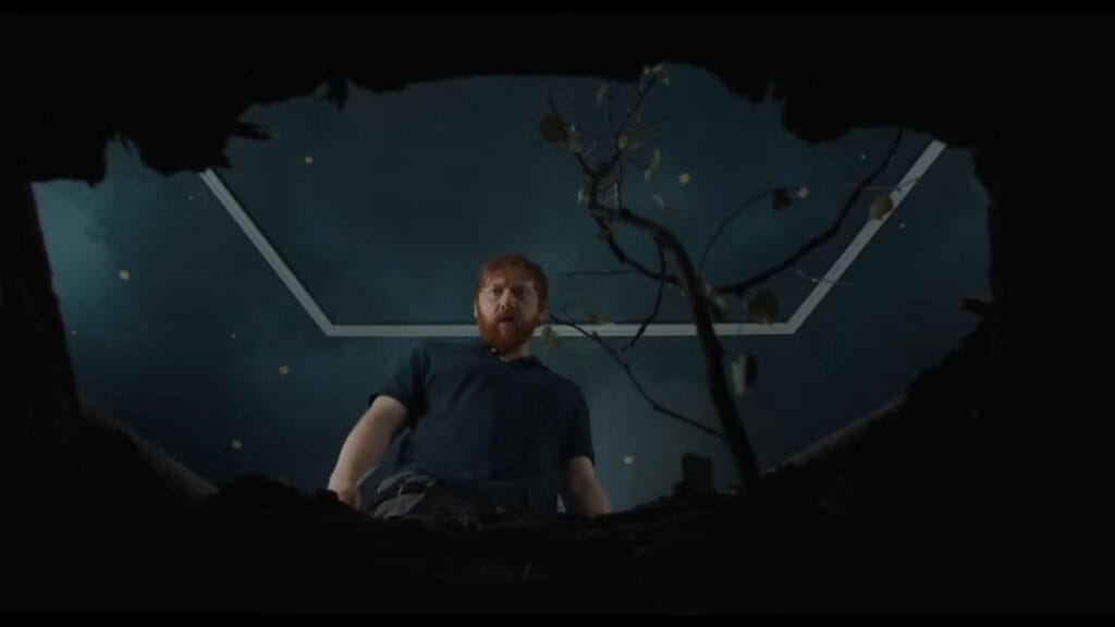A low-angle shot looking up through a hole in the floorboards at a bearded man staring down beside an emerging sapling in Nightborn.