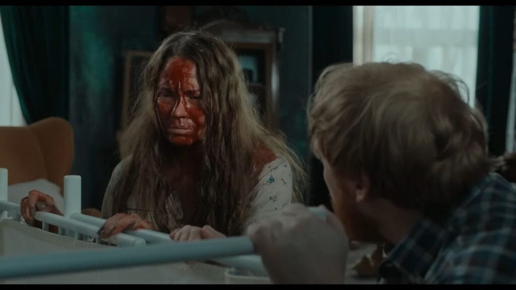 A blood-covered woman gripping a white baby cot and staring intensely at a man in a dark nursery room in Nightborn.