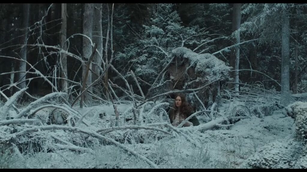 Seidi Haarla as Saga sitting alone amidst fallen, snow-covered tree branches in a dense winter forest in Nightborn.
