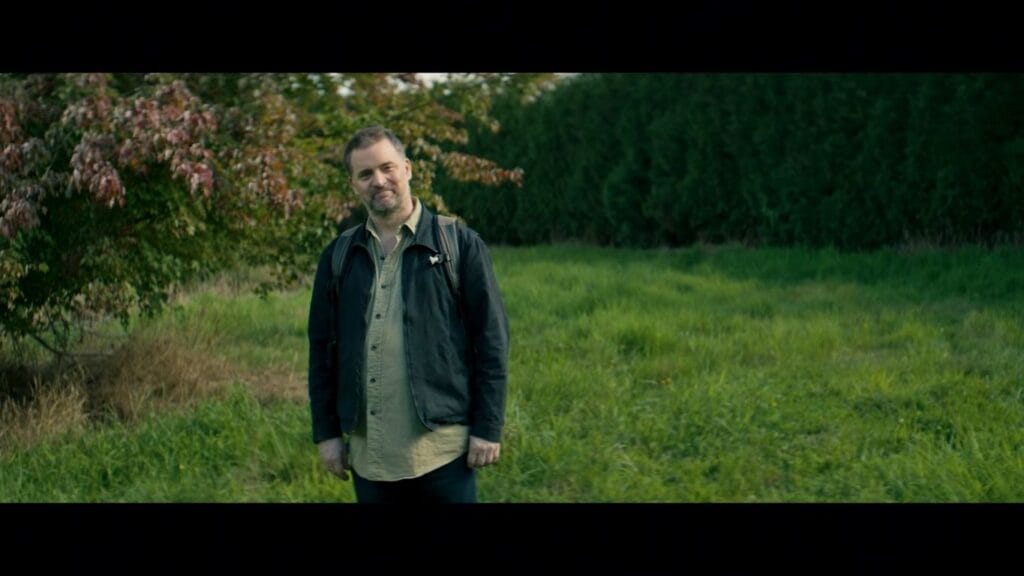 A smiling man wearing a jacket and backpack standing in a green field in Sawtooth Mountain.