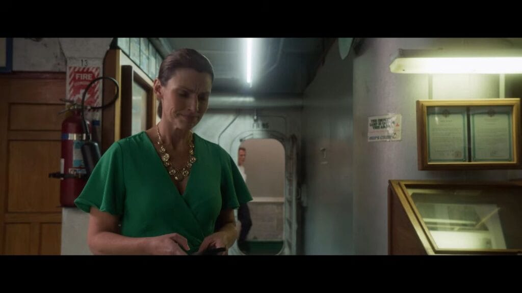 Sarah Lieving as Molly wearing a green dress and gold necklace looking down at her phone in a ship hallway in Shark Terror.