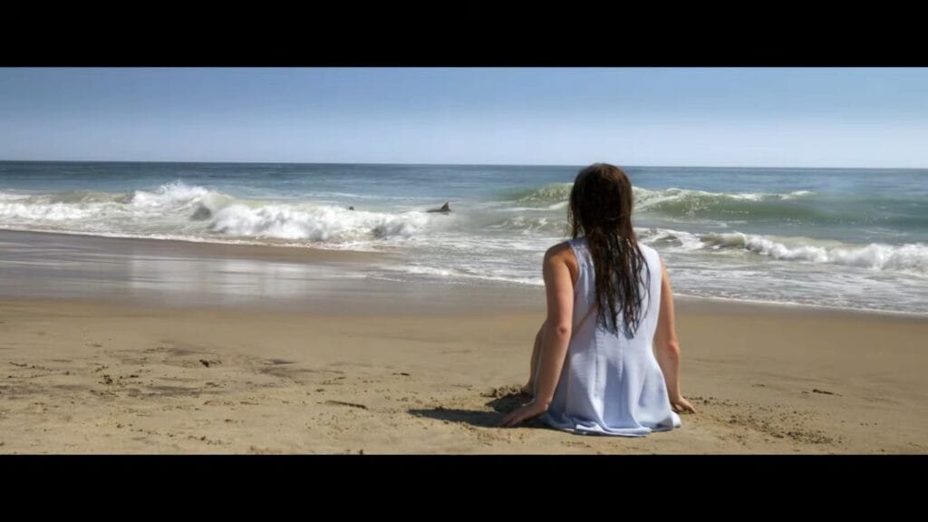A woman in a light blue dress sitting on a sandy beach looking out at ocean waves and a shark fin in Shark Terror.