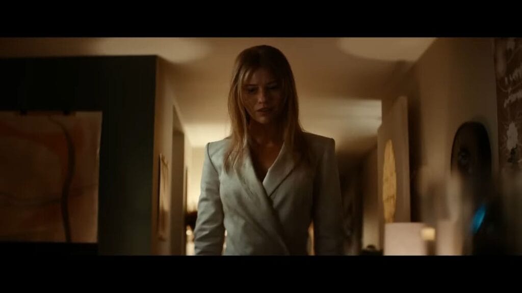 Lily Sullivan as Sara wearing a light grey blazer and looking down while walking through an apartment in Soulm8te.