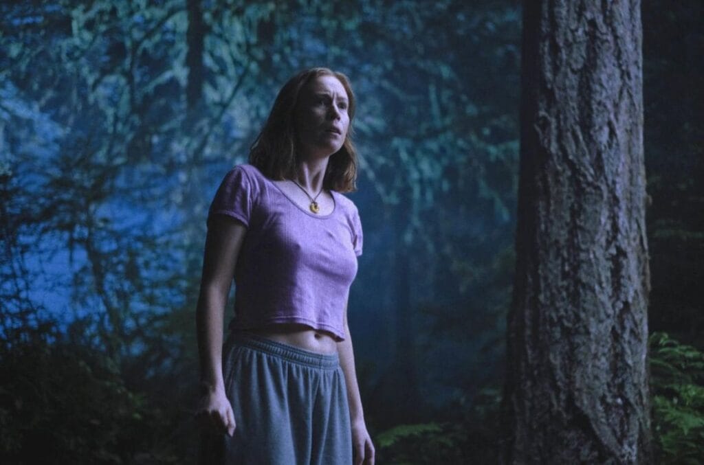A woman in a pink crop top and grey sweatpants looking concerned in a dark, blue-lit forest in Teen Sex and Death at Camp Miasma.