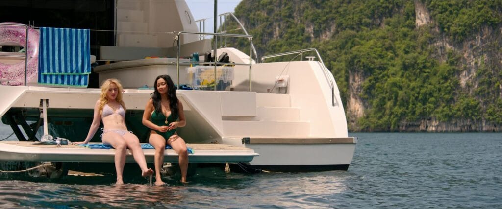 Kathryn Newton and Lana Condor in bikinis sitting on the back ledge of a white yacht dangling their feet in the water in The Devil's Mouth.