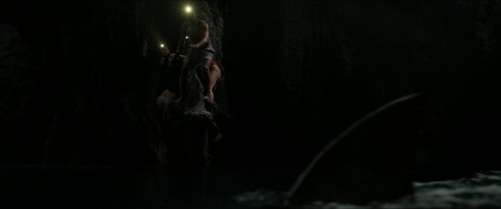 A group of young adults clinging to a dark rock formation inside a cave as a large shark fin circles below in The Devil's Mouth.