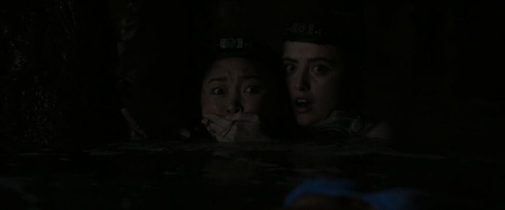 Kathryn Newton covering Lana Condor's mouth as they hide together in fear in a dark cave scene from The Devil's Mouth.