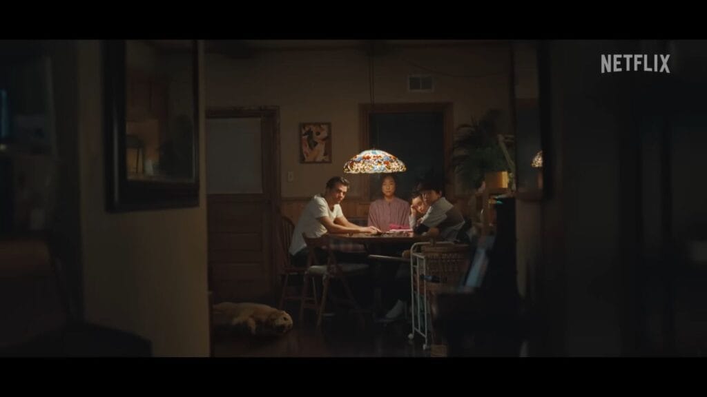 Wagner Moura, Greta Lee, and their children sitting around a dining table under a lamp inside a dark home in The Last House.