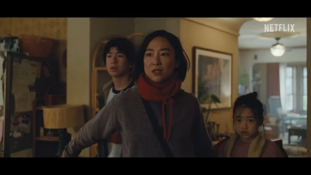 Greta Lee looking alarmed with an outstretched arm while shielding her two children inside a hallway in The Last House.