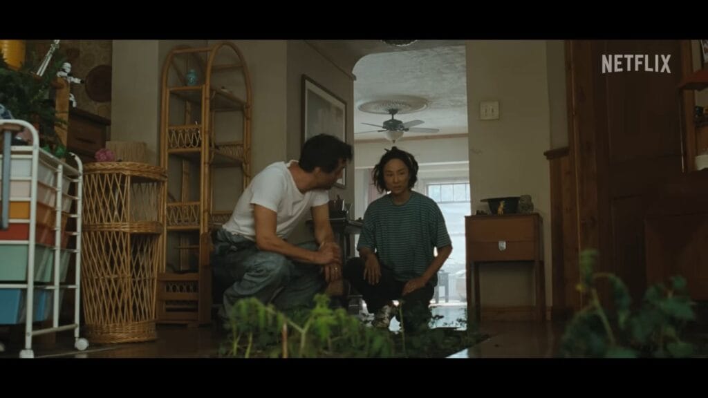 Wagner Moura and Greta Lee crouching down indoors beside indoor potted plants and gardening patches in The Last House.