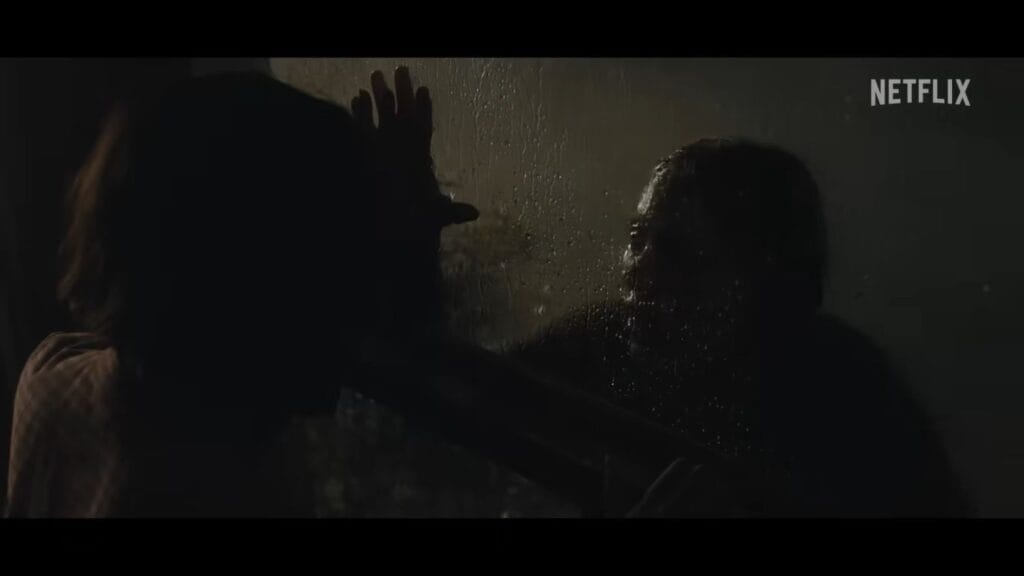 A person pressing their hand against a dark, rain-streaked window while looking at a shadowy figure outside in The Last House.